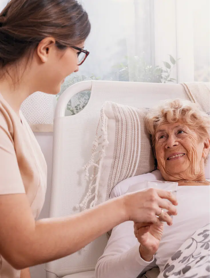 Pflegekraft hält einer älteren Dame die Hand, welche in einem Pflegebett liegt